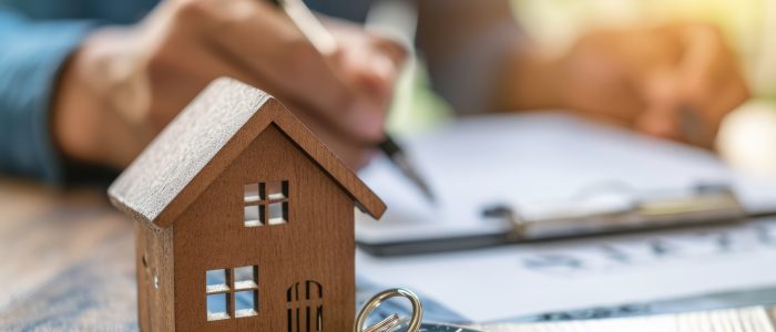 Person unterschreibt Vertrag mit Modellhaus und Schlüsseln im Vordergrund