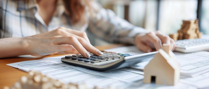 Frau sitzt vor Plänen und rechnet mit einem Taschenrechner ihr Budget aus mit einem Modellhaus im Vordergrund