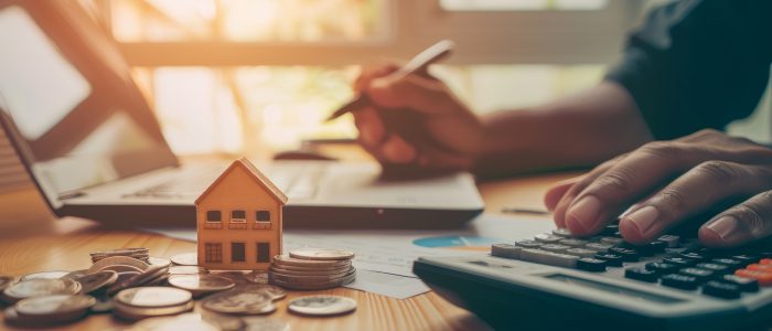 Person am Schreibtisch arbeitet am Laptop und rechnet mit Taschenrechner Kosten aus, Modellhaus und Münzen liegen im Vordergrund