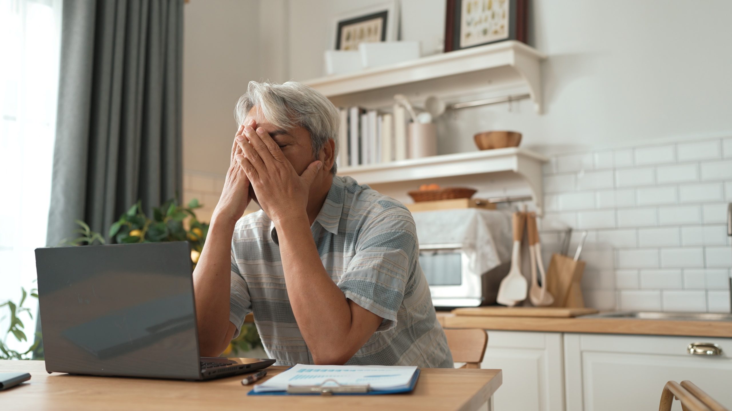 Man sitzt am Computer in der Küche und hält sich die Hände vor sein Gesicht